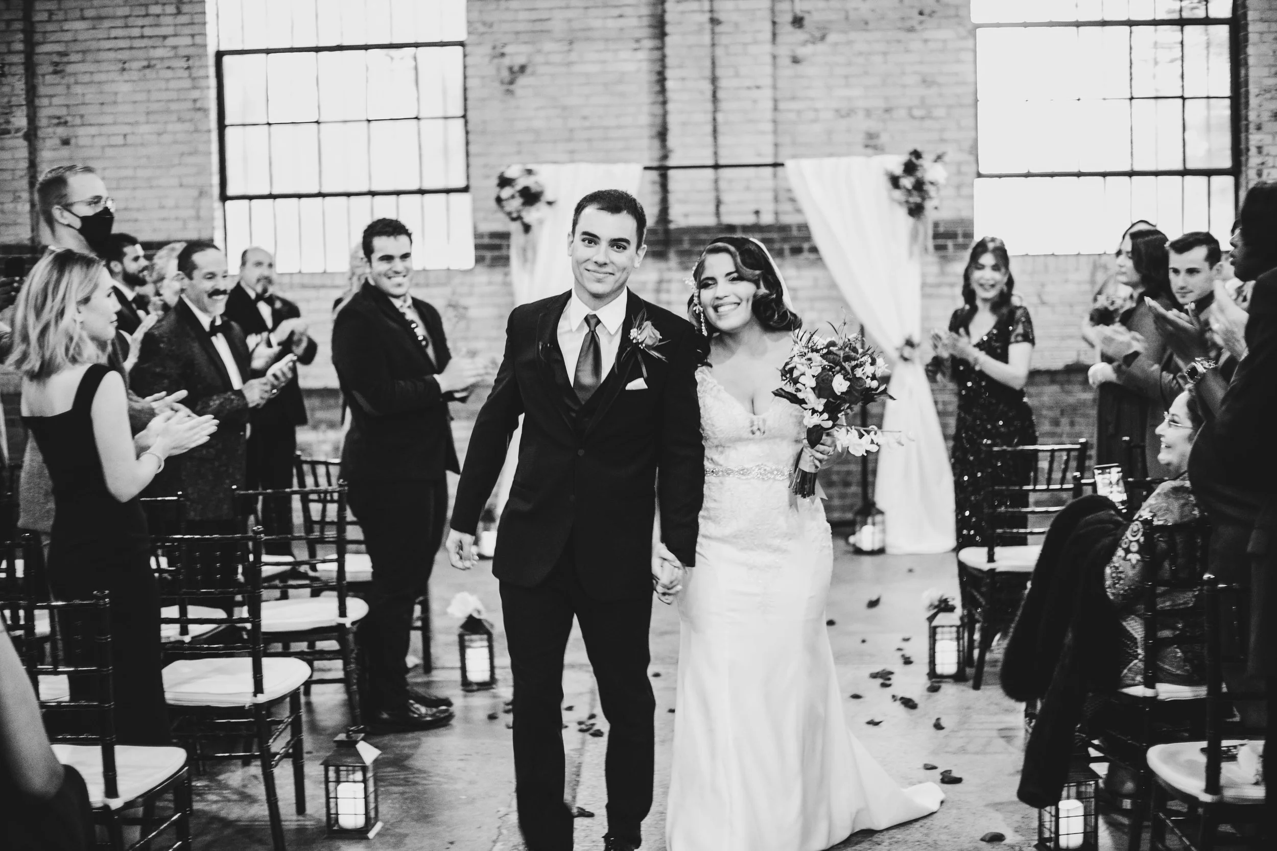A black and white photo of a newlywed couple walking down the aisle, holding hands, surrounded by clapping guests in a brick-walled venue with large windows and floral decorations.