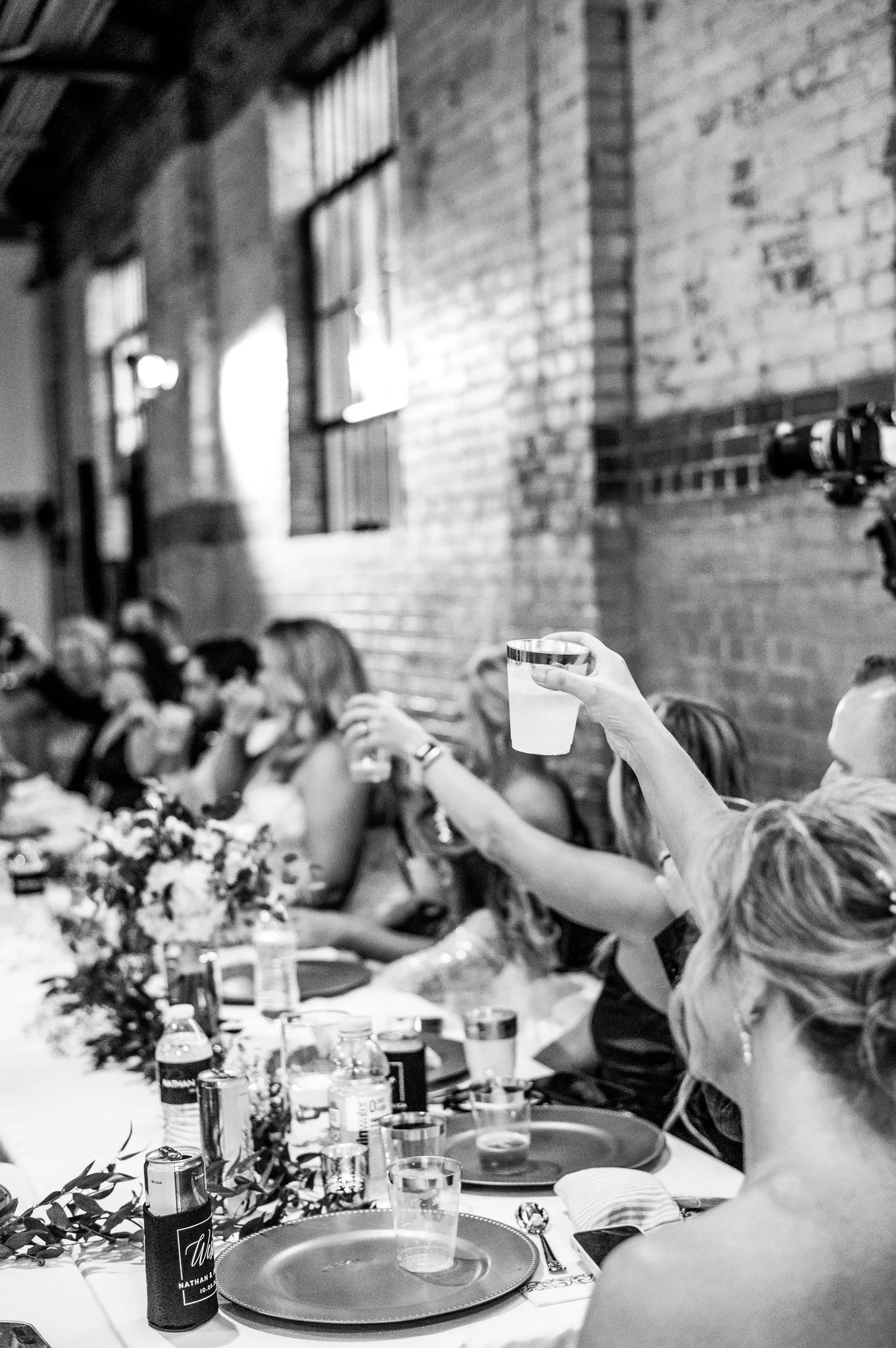 A person raising a drink at a long table with other people, decorated with flowers and candles, in a rustic indoor setting with brick walls and large windows.