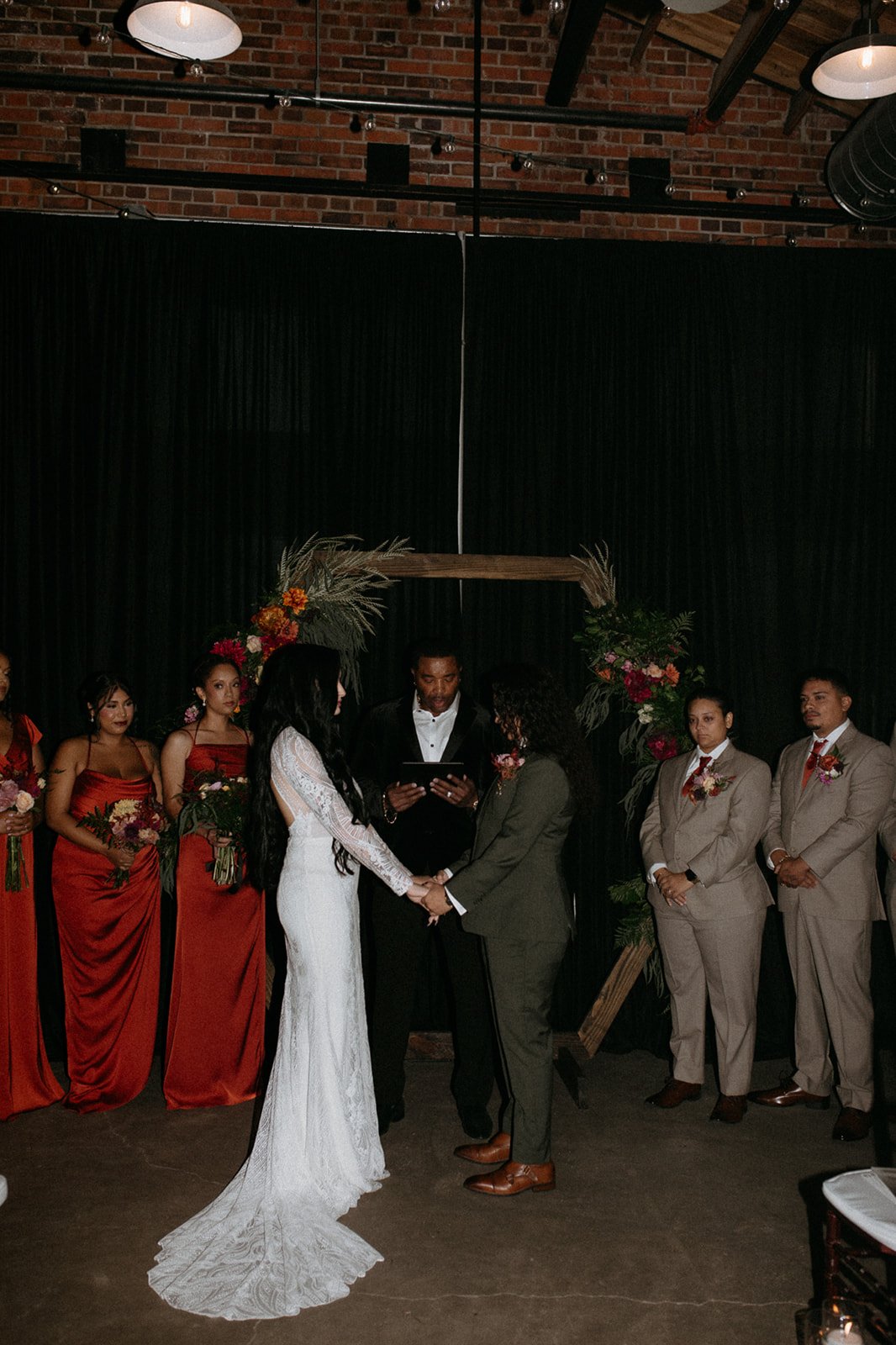 A wedding ceremony with a couple holding hands and facing each other, in front of an officiant, with bridesmaids and groomsmen standing beside them, decorated with floral arrangements and a wooden arch in a brick-walled venue.