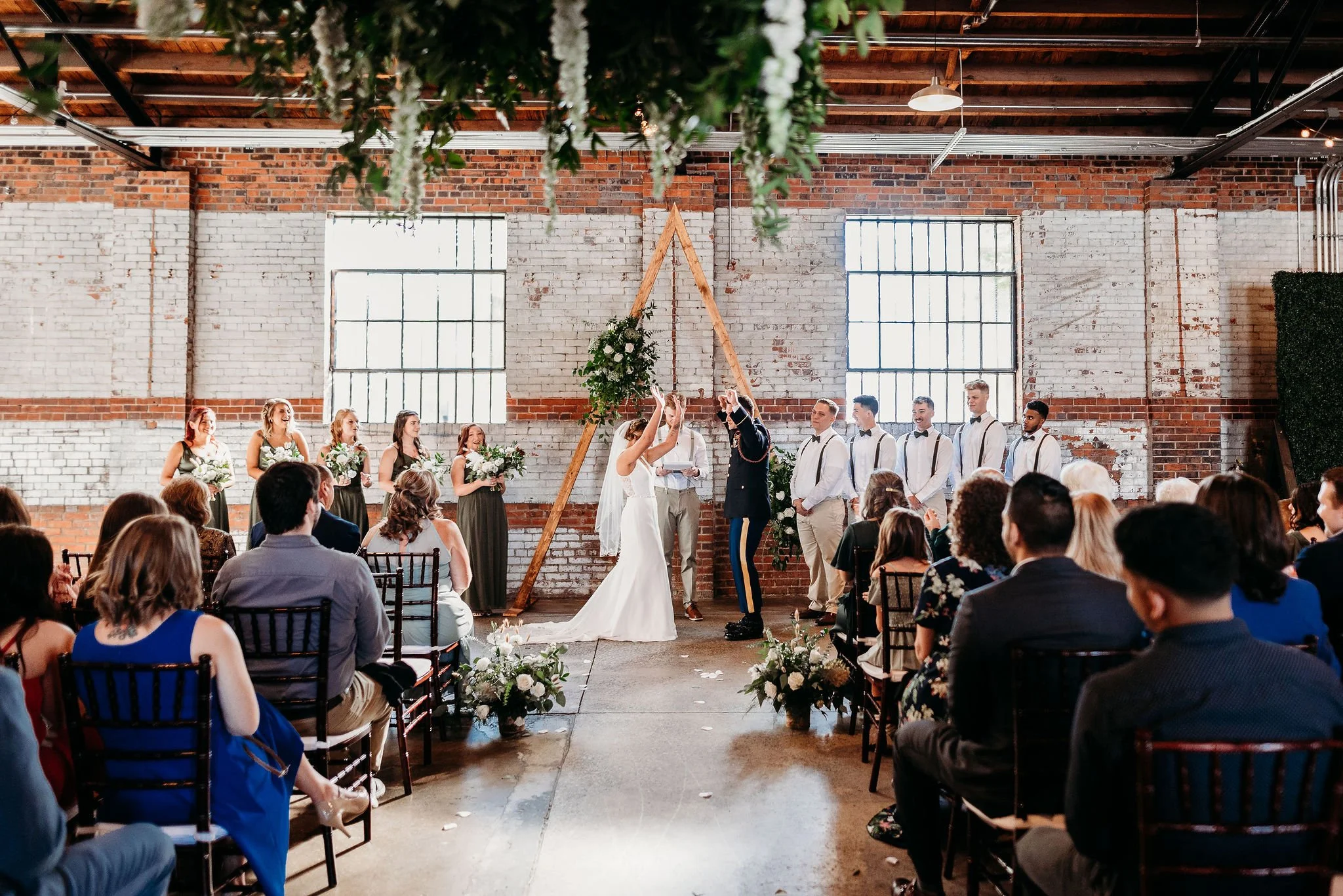 A wedding ceremony taking place inside an industrial-style venue with brick walls and large windows, featuring a bride and groom exchanging vows in front of their wedding party and seated guests.