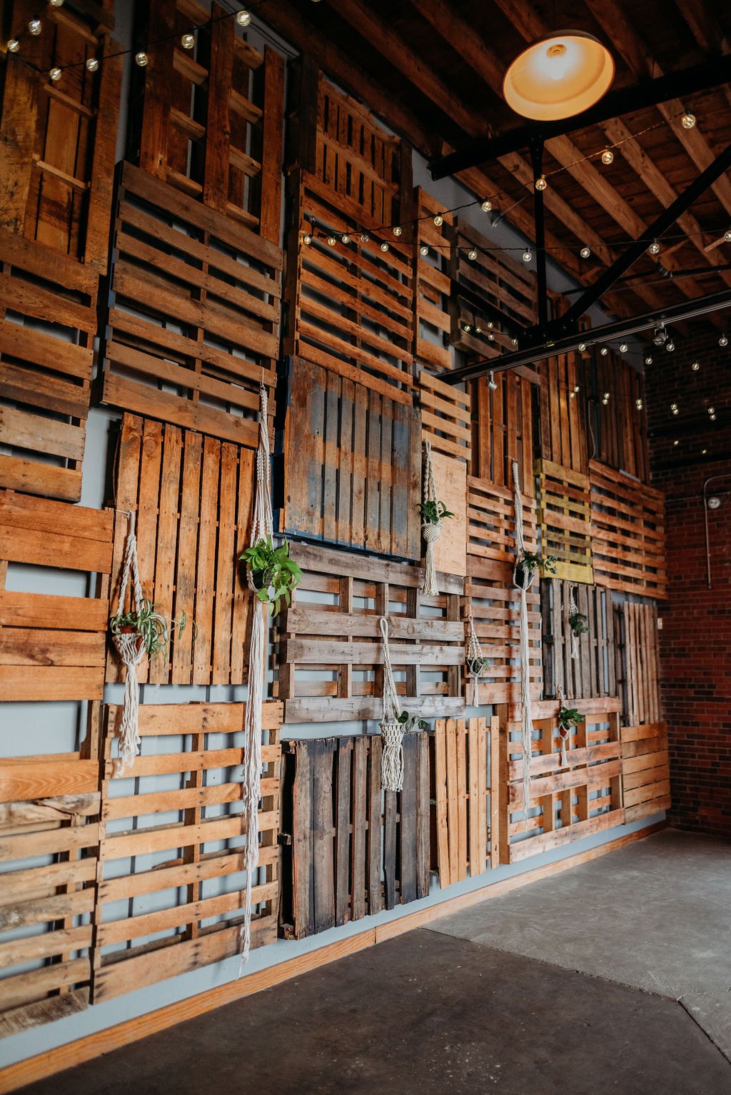 Decorative wall made of wooden pallets with hanging plants in macrame holders and string lights, in an industrial-style interior.