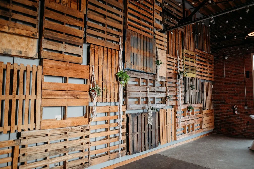 Decorative wall made of wooden pallets and planks arranged in a pattern, with hanging plants and string lights in a modern interior space.