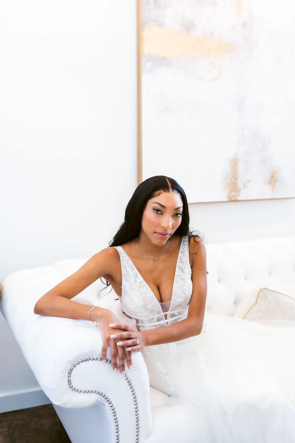 A woman with long black hair sitting on a white sofa in a bright room, wearing a white lace dress with a deep neckline, looking at the camera.