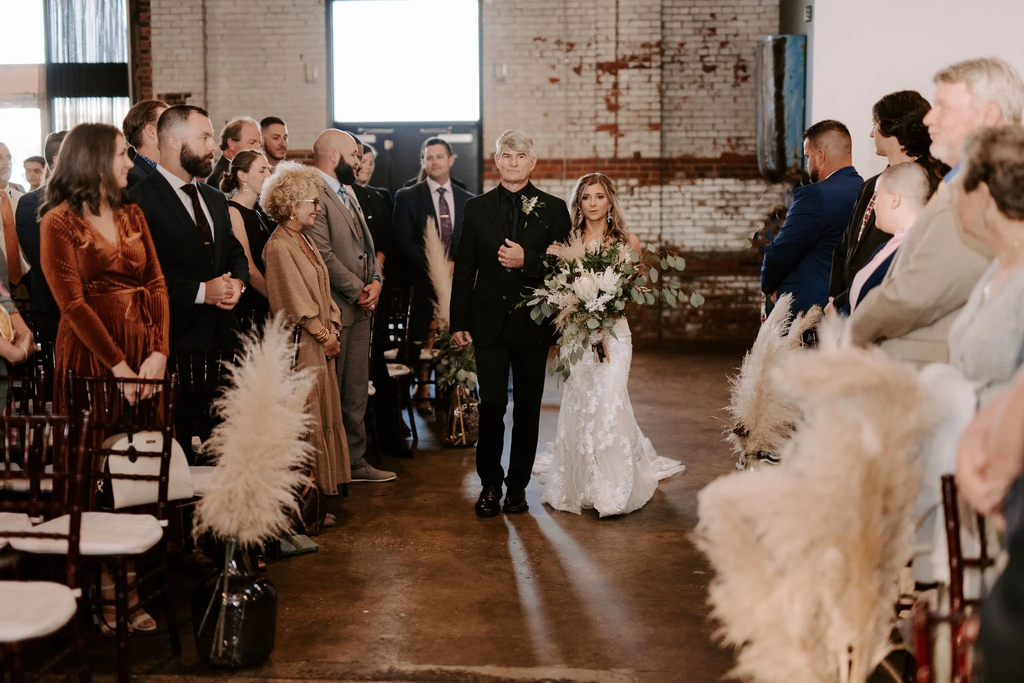 Bride walking down the aisle escorted by a man at her wedding ceremony, with guests standing on both sides of the aisle in an industrial-style venue with exposed brick walls.