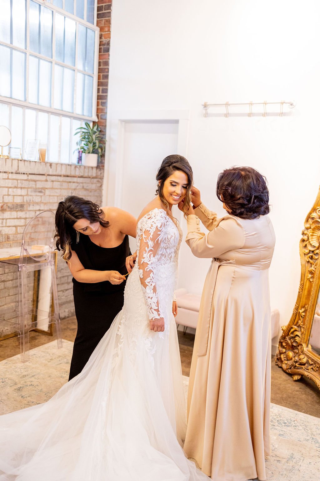 A bride in a white wedding gown is being assisted by two women, one adjusting the dress and the other helping with earrings, in a bright room with a large mirror and brick wall.