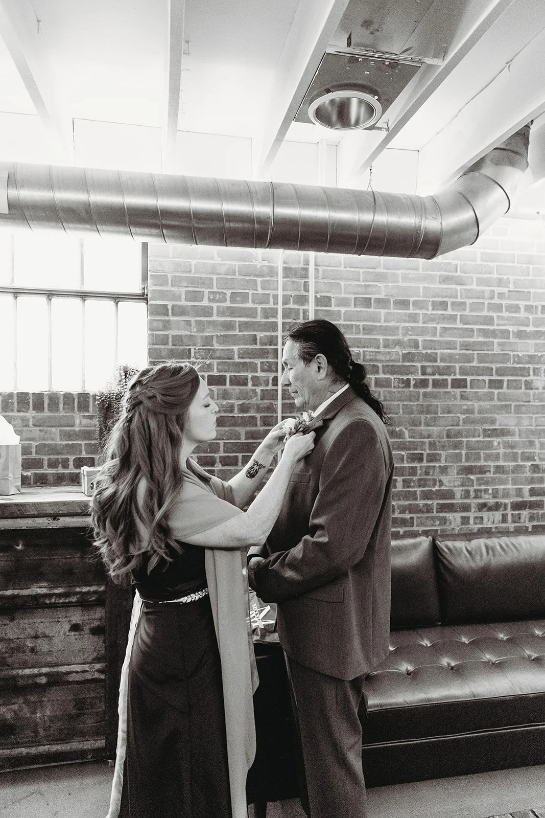 A woman helps a man adjust his bowtie in a room with brick walls and industrial ceiling ductwork.