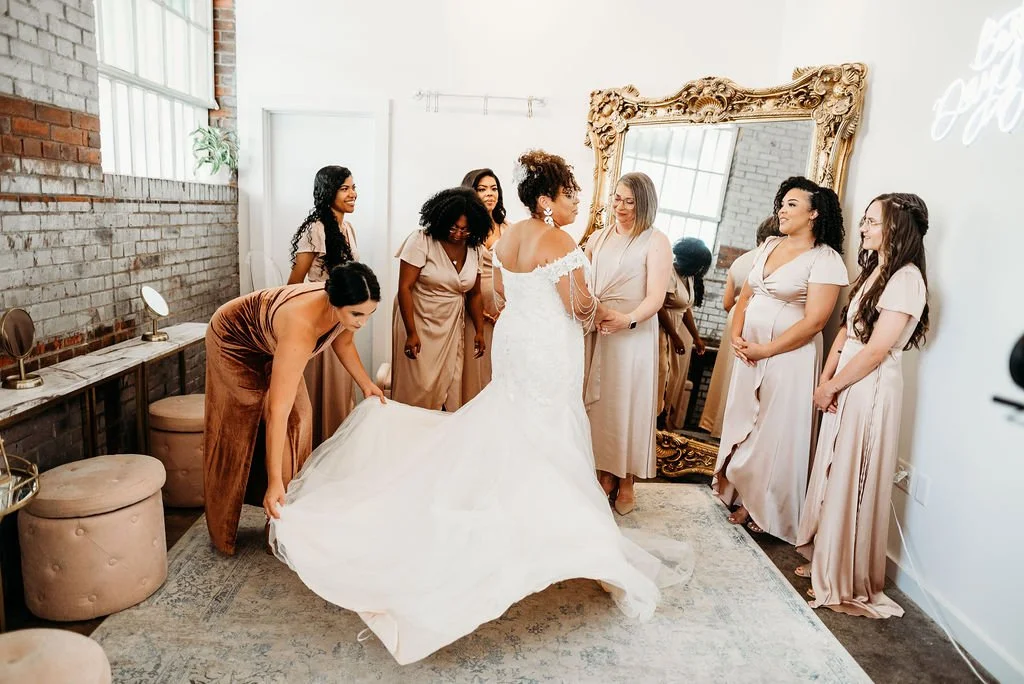 Bride in a white wedding dress surrounded by her bridesmaids in matching dresses, preparing for the wedding in a bright room with a large ornate mirror and exposed brick wall.