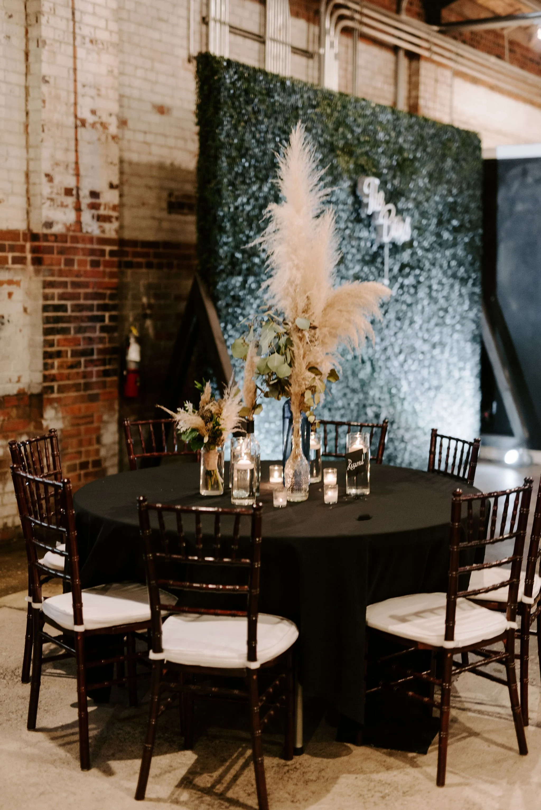 A round table decorated with tall feather floral centerpieces and candles, set for an event inside a brick-walled venue.
