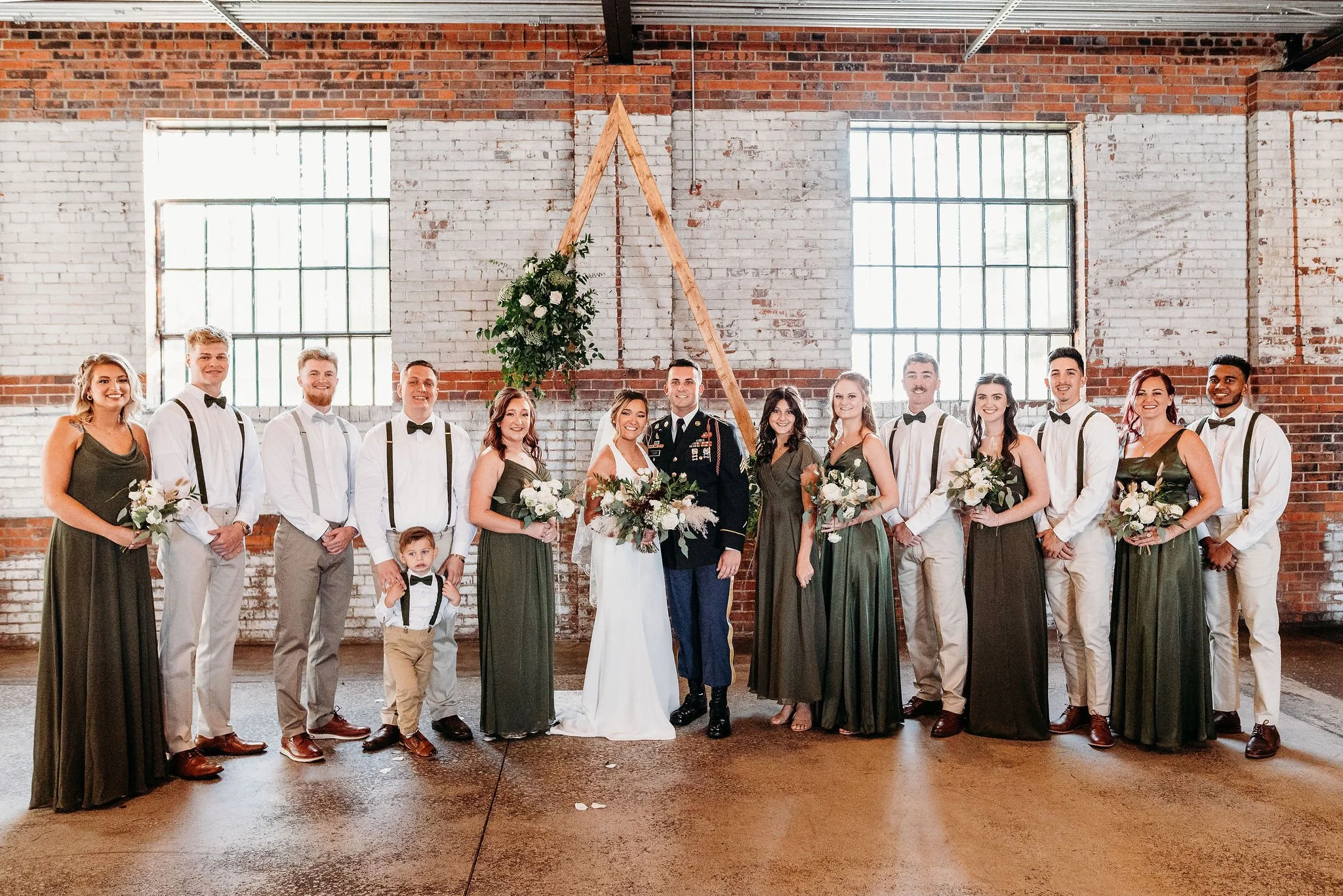 Bride and groom with wedding party in a rustic indoor setting with brick walls and large windows, including bridesmaids and groomsmen, some holding bouquets, and a young boy in suspenders and bowtie.