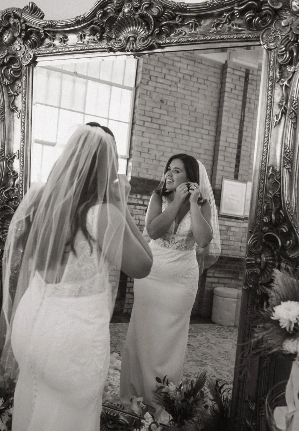 A bride is standing in front of an ornate mirror, adjusting her earring while smiling. She is wearing a white wedding dress and a veil. The setting appears to be a rustic indoor space with brick walls and large windows, decorated with flowers.