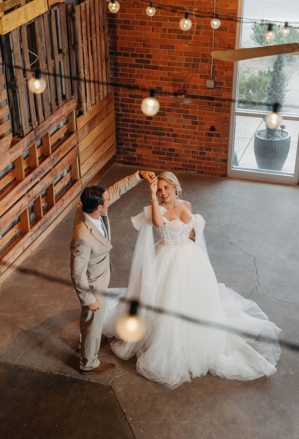 A bride and groom dancing inside a rustic venue with exposed brick walls and a large window. The bride is wearing a white wedding gown and the groom a beige suit. String lights hang from the ceiling, creating a warm atmosphere.