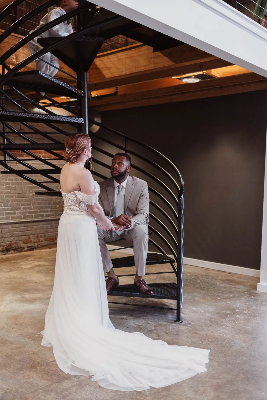 A bride and groom are having a moment beneath a spiral staircase indoors. The bride, in a white lace off-shoulder wedding dress with a long train, stands facing the groom, who is seated on a black metal chair, wearing a light gray suit and tie. There