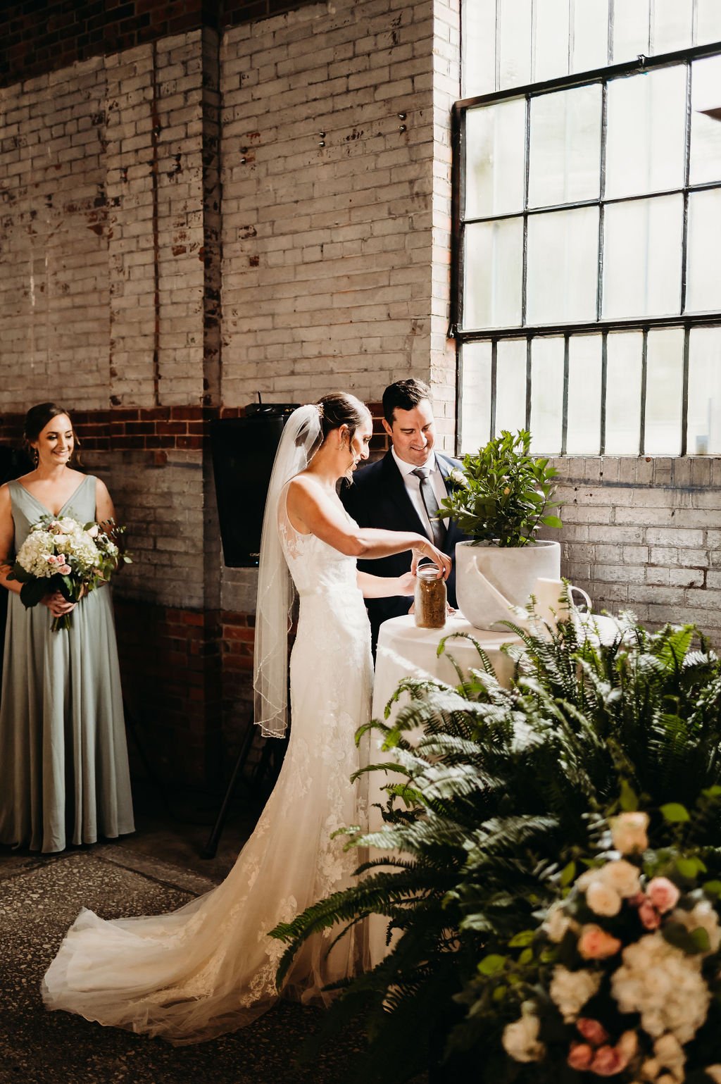 A bride and groom exchanging vows at their wedding ceremony next to a large window with a leek plant and flowers nearby, with a bridesmaid holding a bouquet in the background.