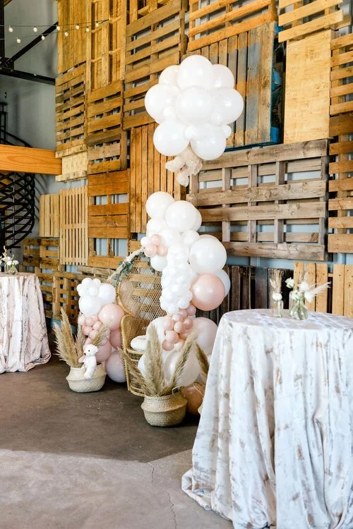 Decorative setup with pink and white balloons, a wicker chair, and a table with a floral arrangement, against a rustic wooden pallet wall.