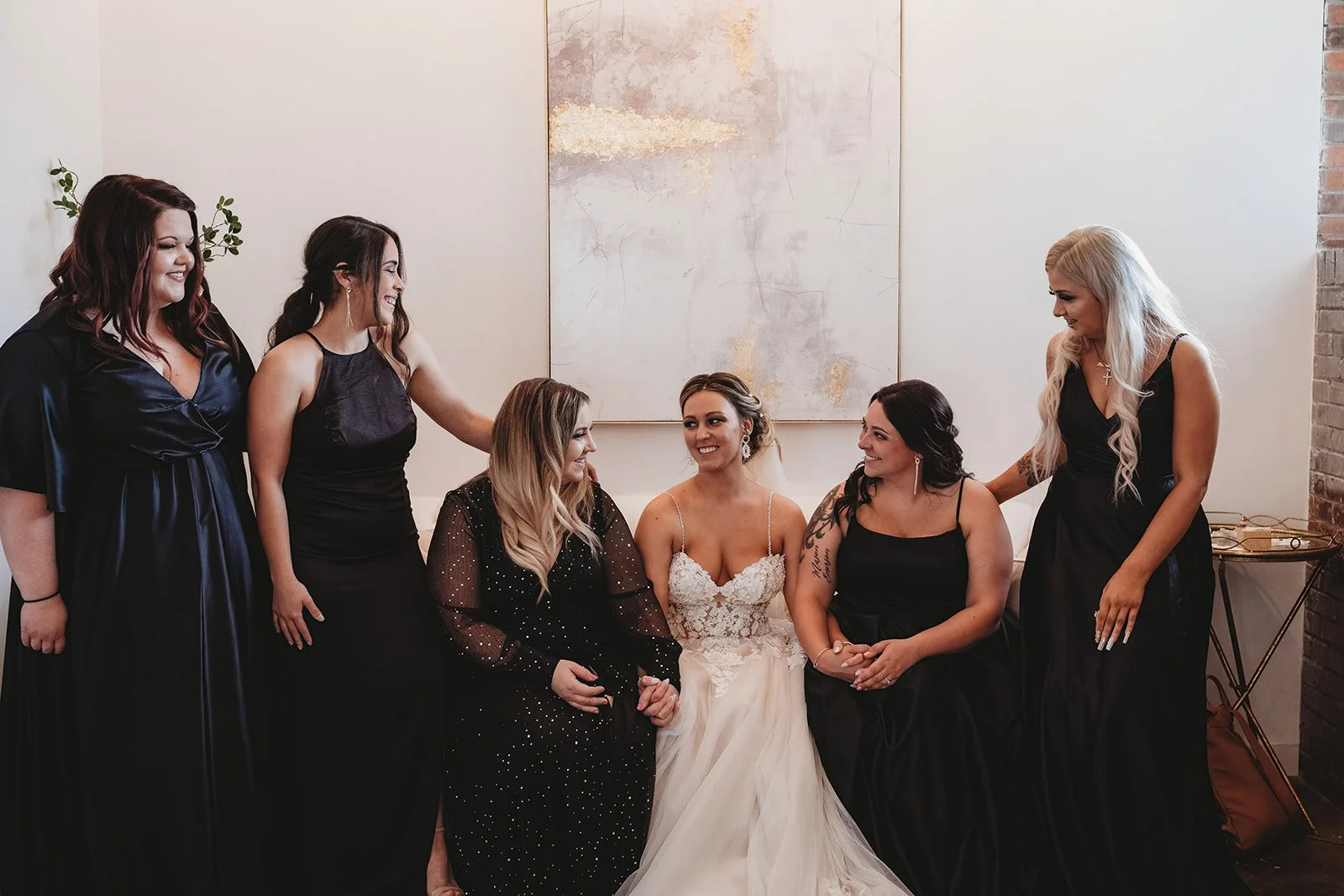A bride in a white wedding dress surrounded by five women in black dresses, all smiling and sitting together inside a room with abstract wall art and brick accents.