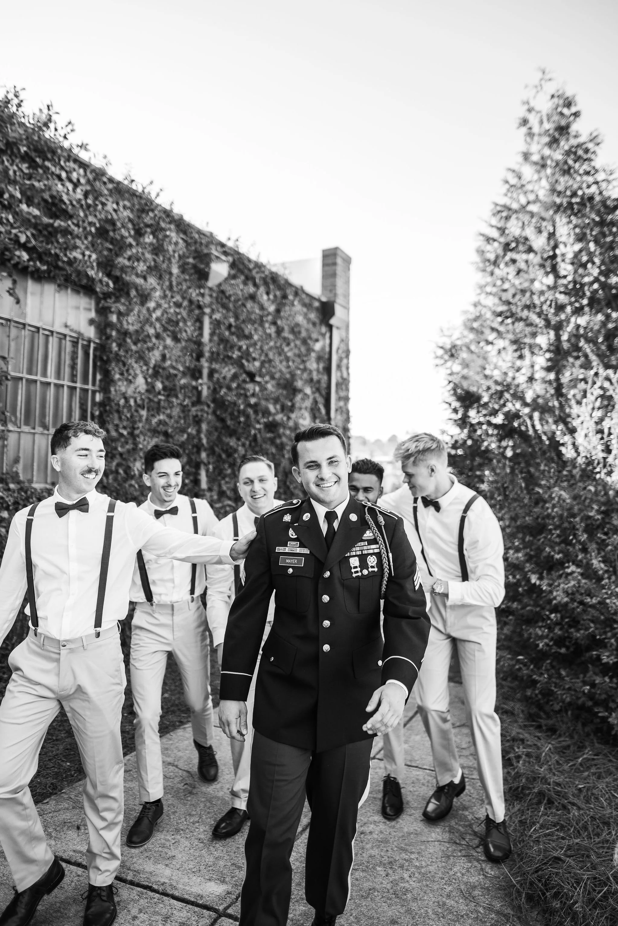 A group of men, with one dressed in a military uniform, walking outdoors on a sidewalk with trees and a building covered in ivy in the background. He is smiling, as are the others.