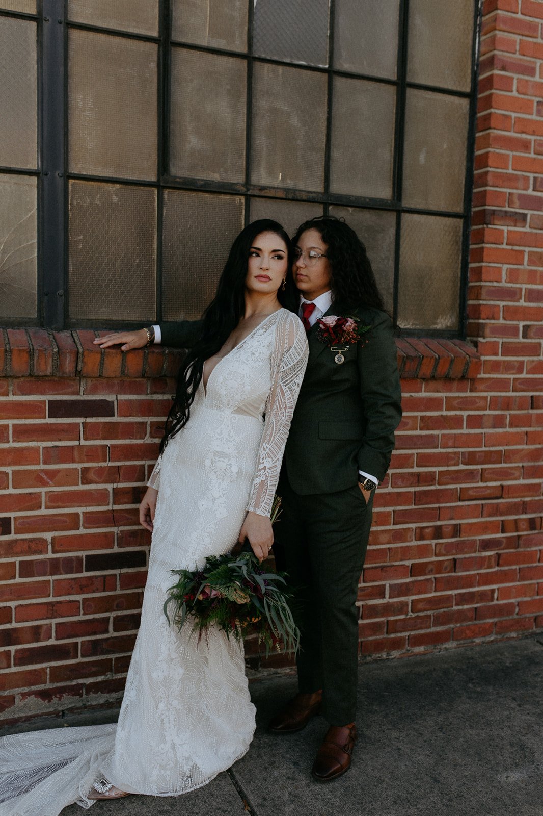 Two women standing against a brick wall, one wearing a white wedding dress holding a bouquet, the other dressed in a dark green suit with a red tie and flower boutonniere, sharing an intimate moment.