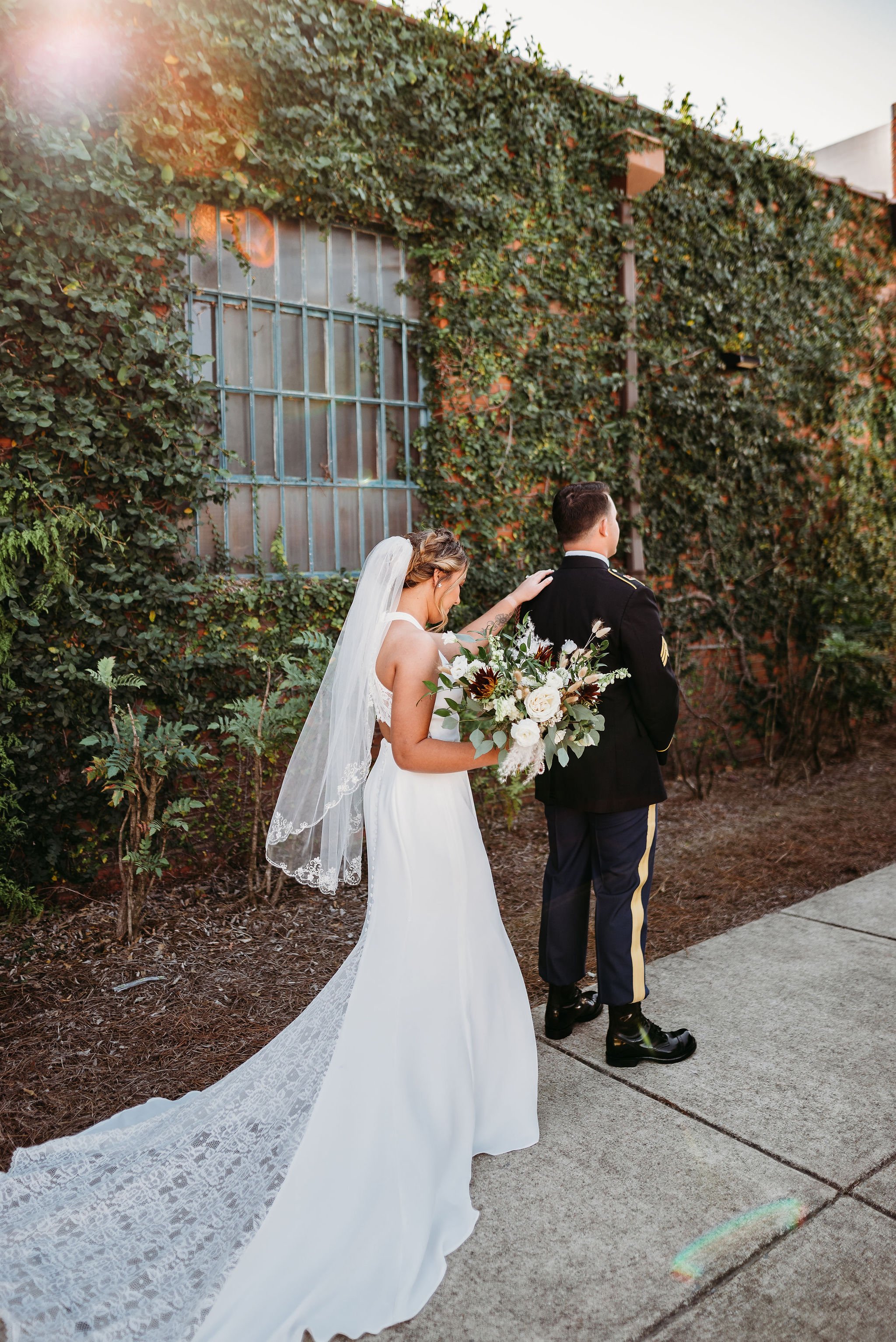 A bride with a veil and white dress, holding a bouquet of flowers, is gently touching a groom in a military uniform, standing outdoors against a brick wall covered in ivy.