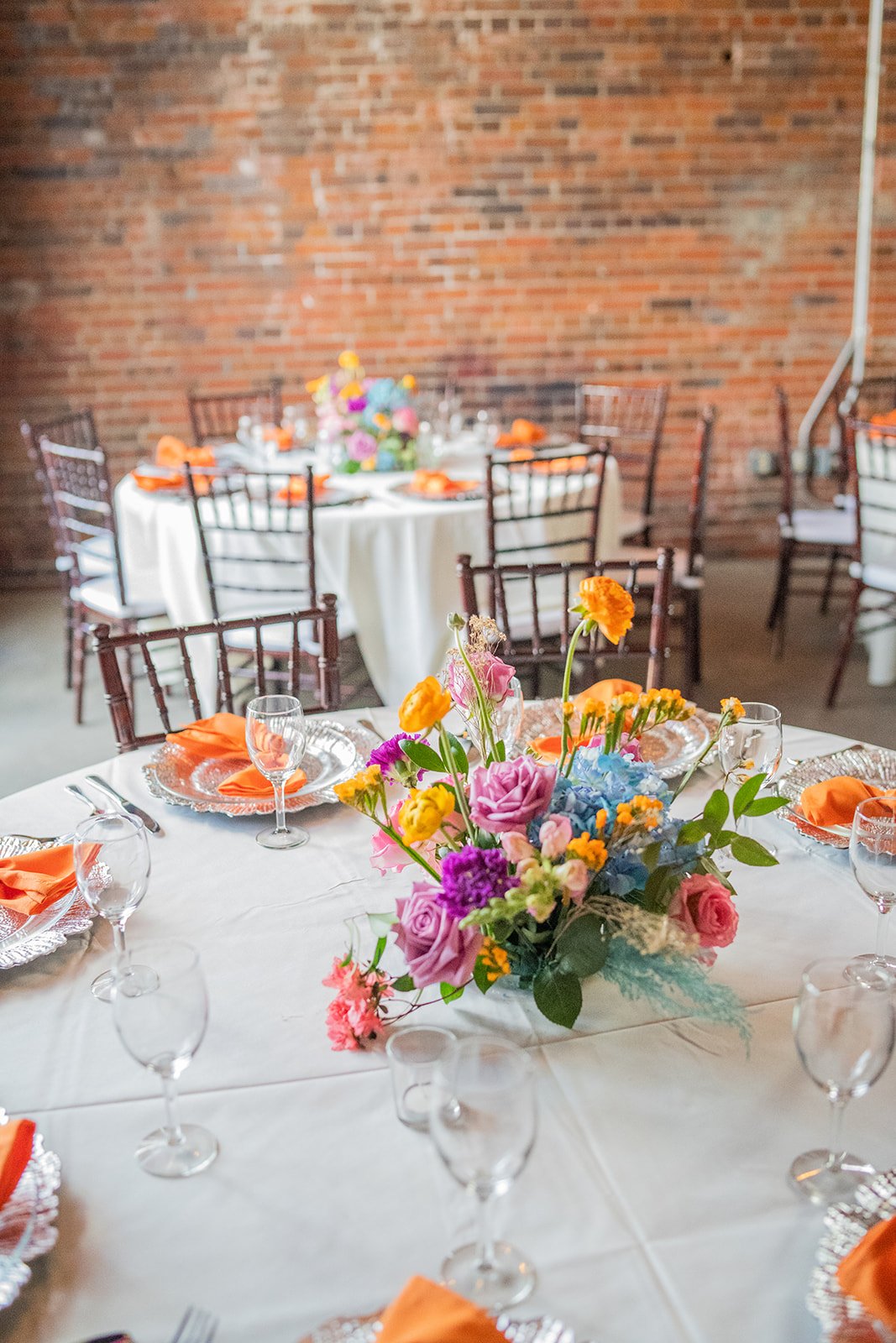 A decorated dining table with a floral centerpiece, surrounded by chairs with orange napkins, in front of a brick wall.