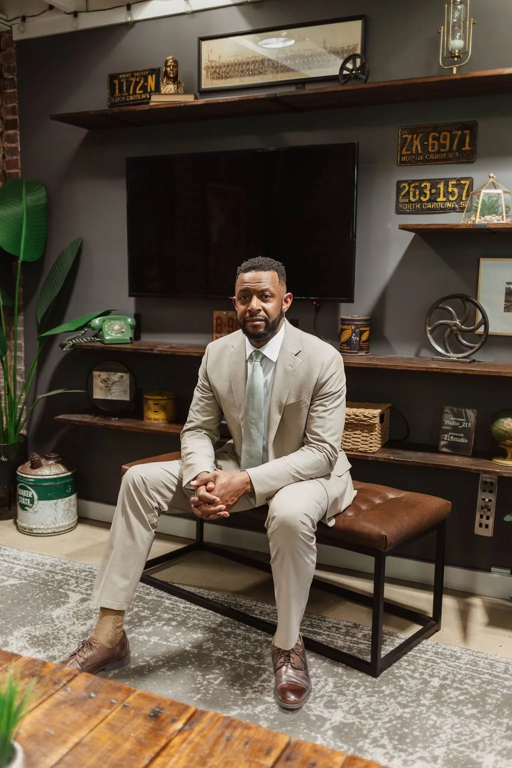 A man in a beige suit and tie sitting on a brown bench in a room with dark gray walls decorated with vintage license plates, a large television, shelves with decorative items, and a large green plant.