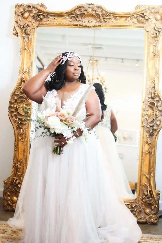 A bride looking at her reflection in an ornate gold mirror, wearing a white wedding dress, holding a bouquet of pink and white flowers, and adjusting her headpiece.
