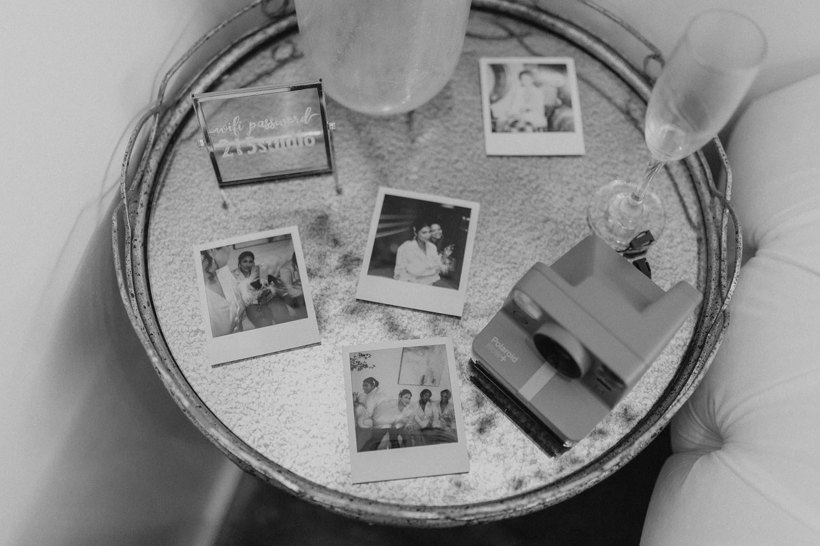 A round metallic table with four Polaroid photos, a Polaroid camera, a champagne glass, a tall glass, a lit candle, and a small sign that reads 'wolf password' in cursive. The table is beside a white cushioned chair.