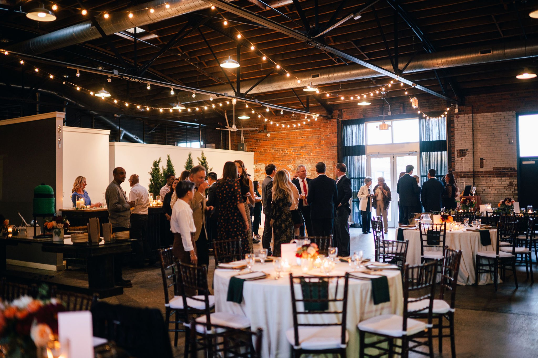 People socializing at a wedding reception or party in a decorated indoor venue with string lights, round tables with white tablecloths, and floral centerpieces.