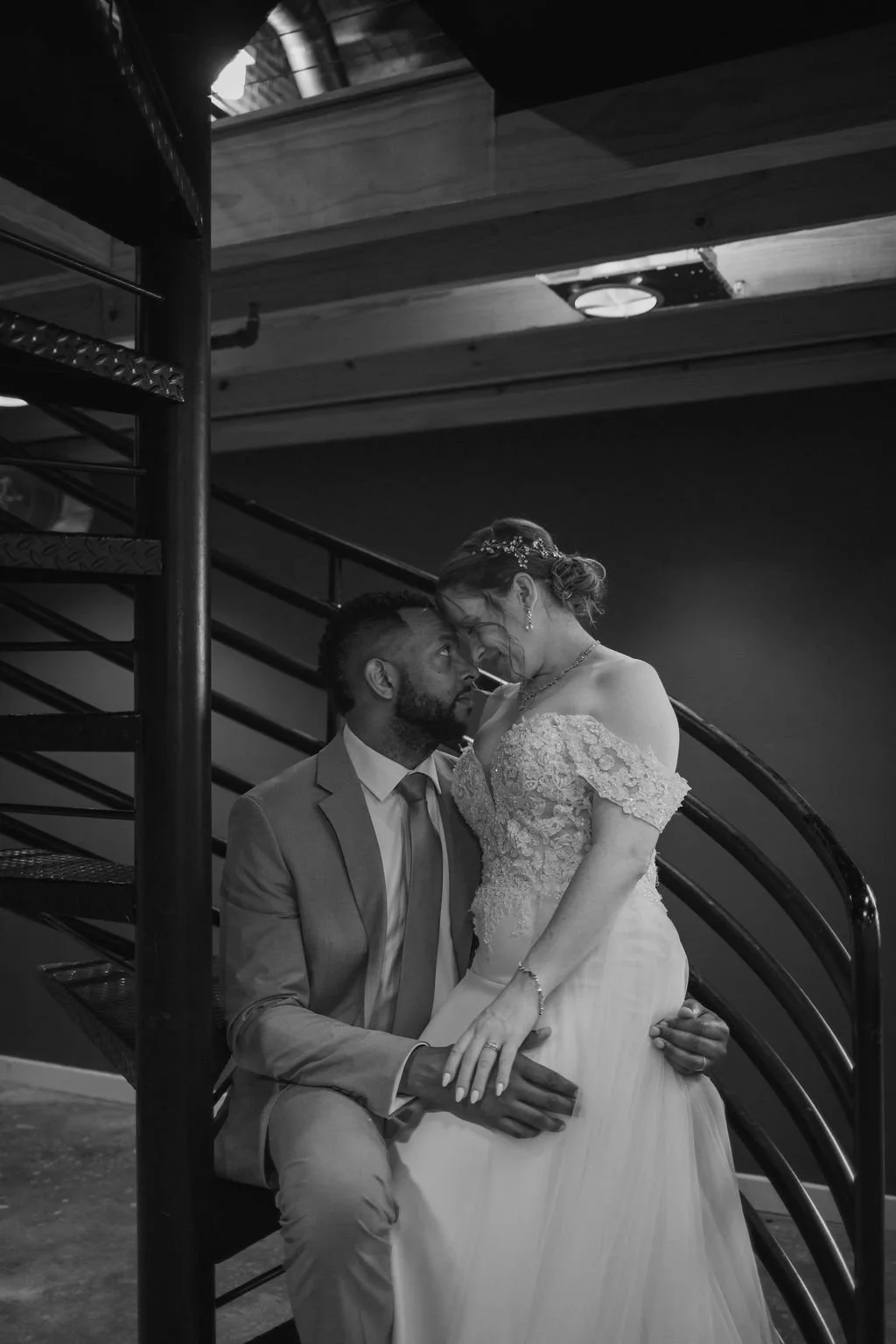 A couple on their wedding day, the groom kneeling while the bride stands, both gazing and touching foreheads, on a staircase in a dimly lit indoor setting.