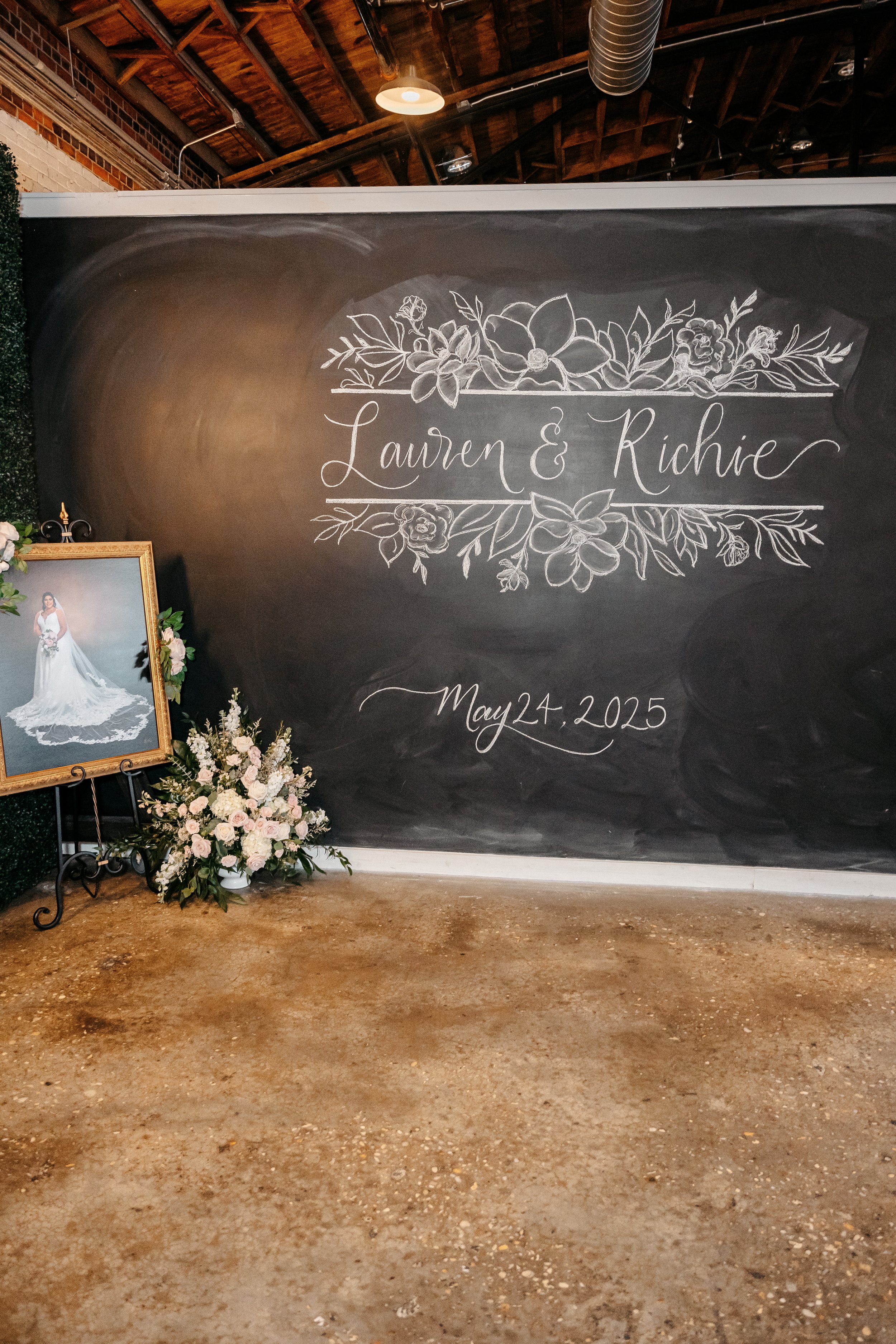 Chalkboard with floral decoration and the names Lauren and Richie, along with the date May 24, 2025, at a wedding event. There is a floral arrangement and a framed photograph of a bride in a wedding dress beside the chalkboard.