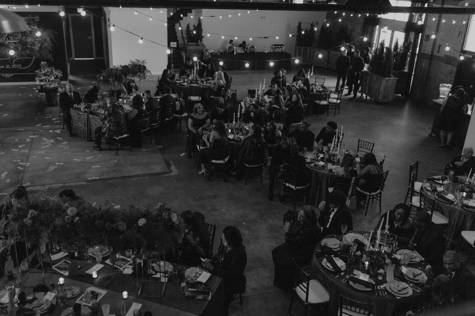 A black and white photograph of a large indoor event with multiple round tables set for a celebration, candle centerpieces, and guests seated and standing around. String lights hang from the ceiling, creating a festive atmosphere.