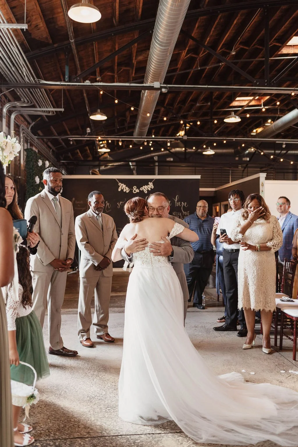 A bride and an older man are hugging in the center of a wedding reception. Guests are standing and watching, some holding phones and emotional. The setting has warm lighting and a rustic, indoor venue with string lights overhead at Studio 215 venue