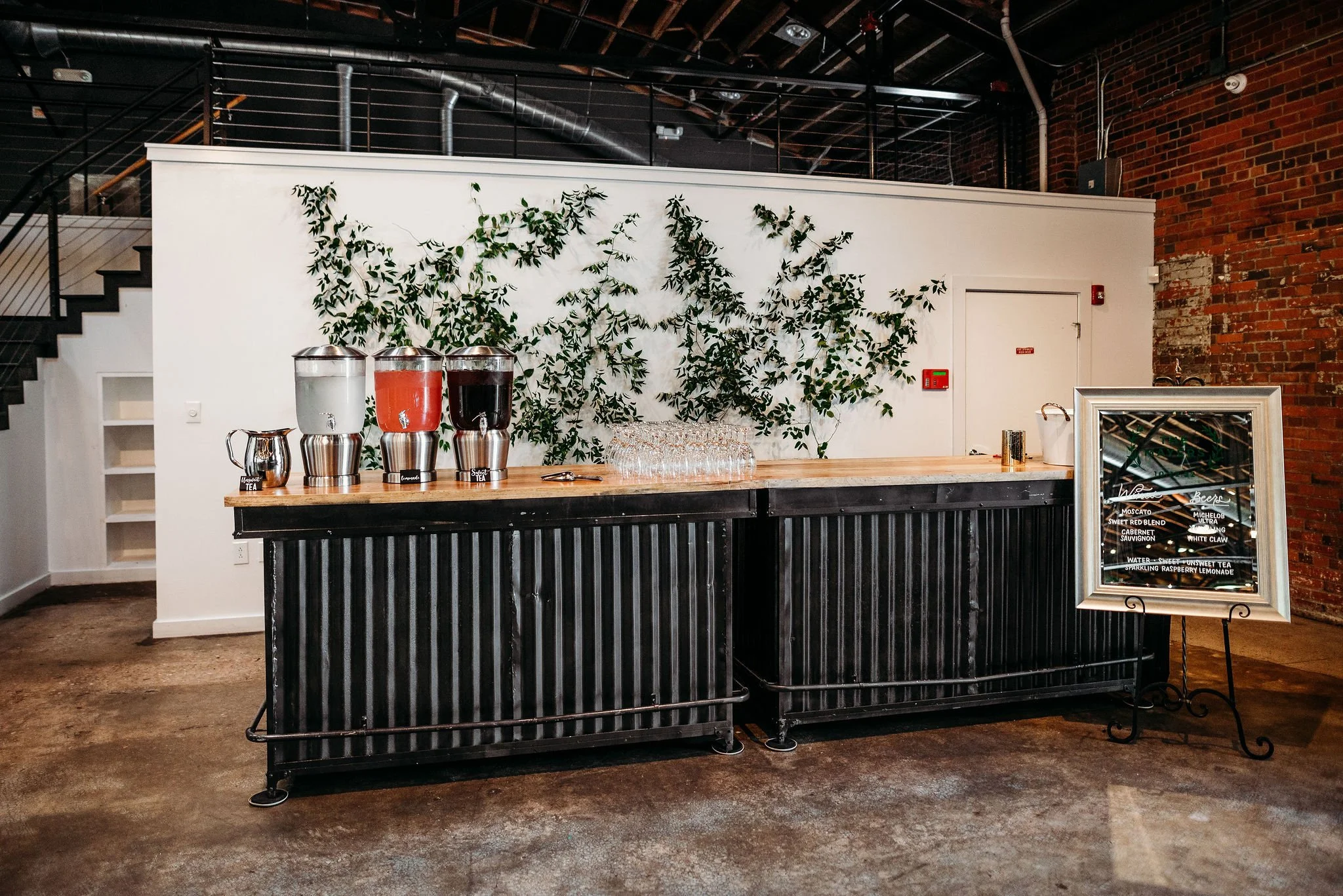 Bar area with three beverage dispensers filled with water, red drink, and dark drink. Glasses stacked nearby, and a framed menu on a stand to the right. Green plant backdrop on the white wall behind the bar, exposed brick wall on the right, and stair