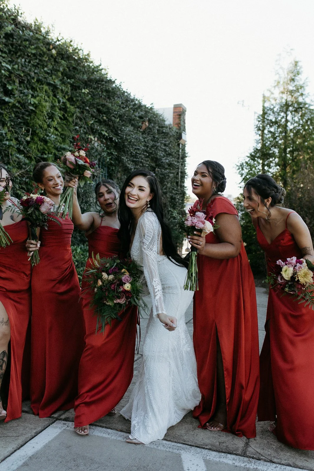 A bride in a white dress with black hair holding a bouquet, standing among bridesmaids in red dresses, smiling and laughing outdoor on a sidewalk with greenery and trees in the background.