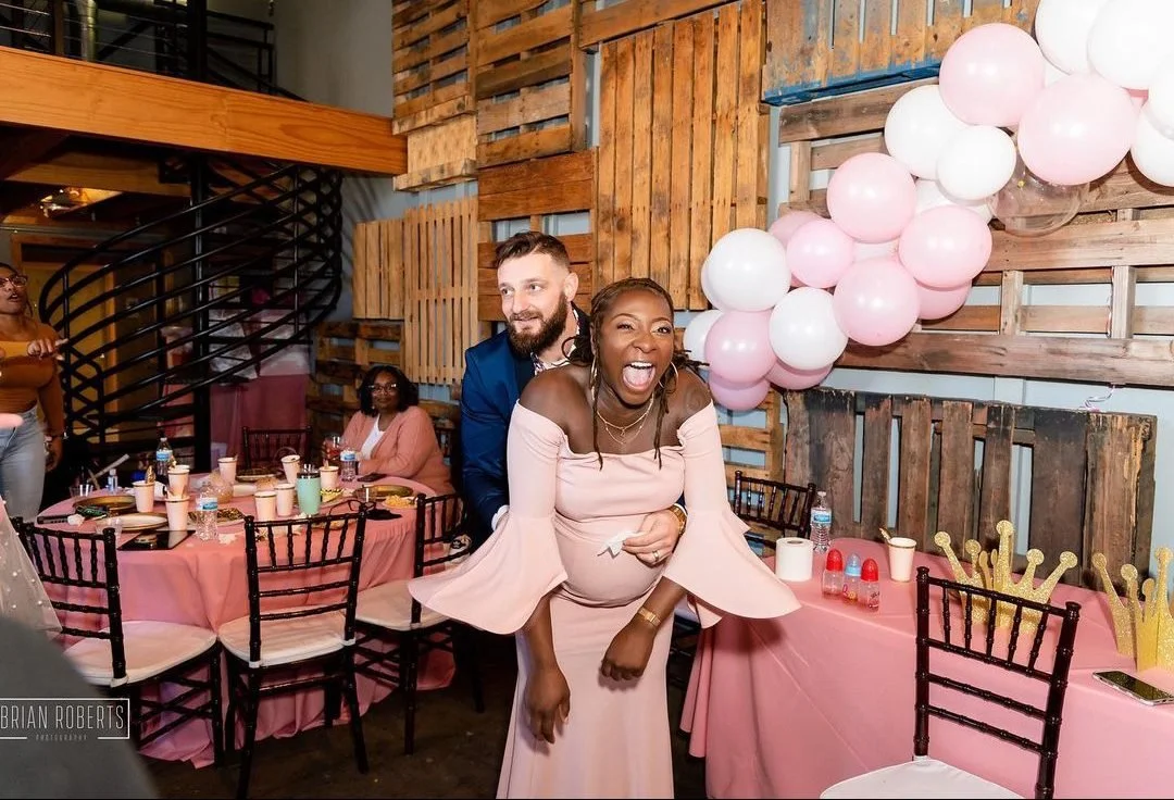A group of people celebrating at a party with pink themed decorations, including pink balloons, a pink tablecloth, and a woman in a pink dress laughing while hugging a man from behind.