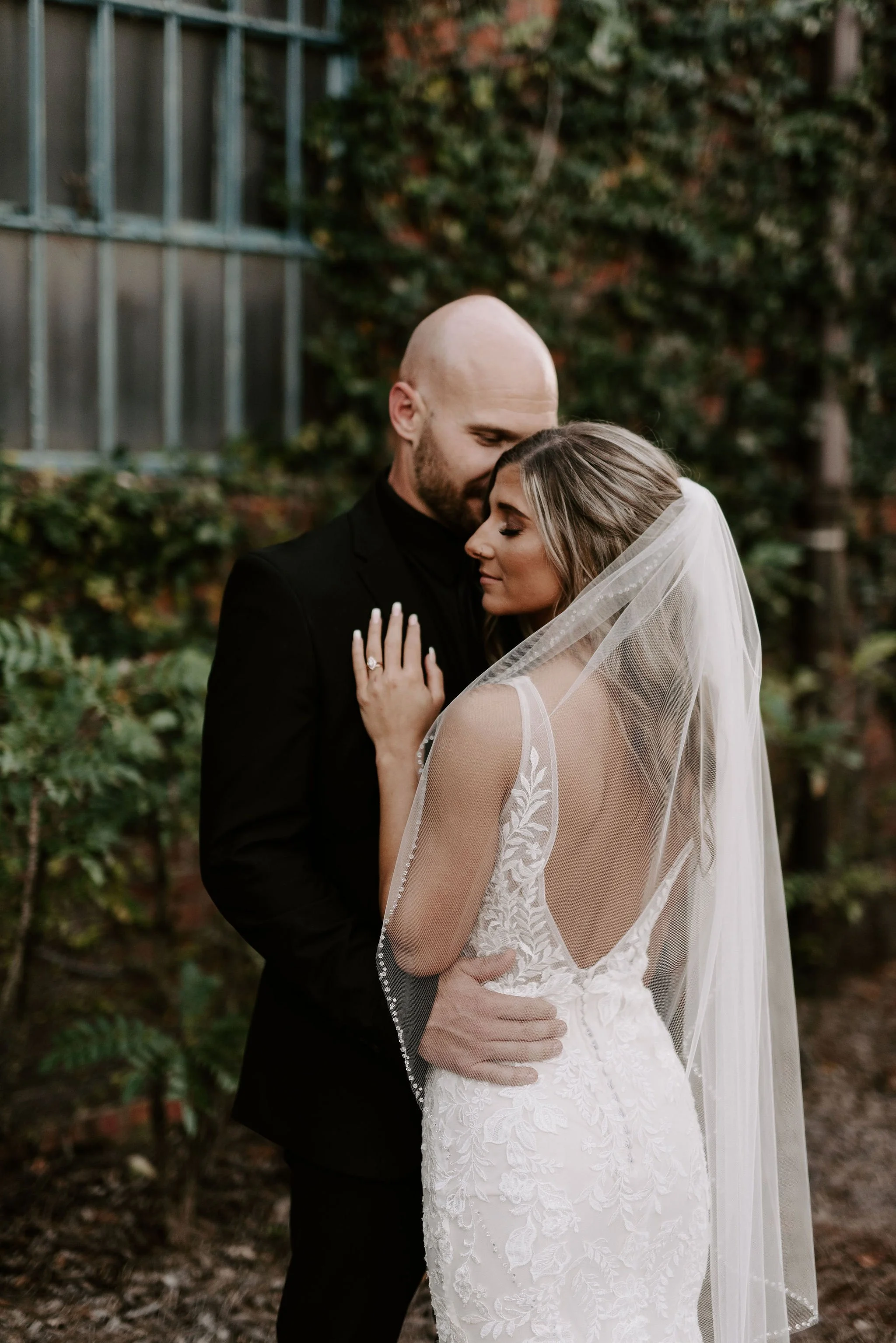 A bride and groom embrace outdoors, with the groom in a black suit and the bride in a lace wedding dress and veil, standing close with their eyes closed.