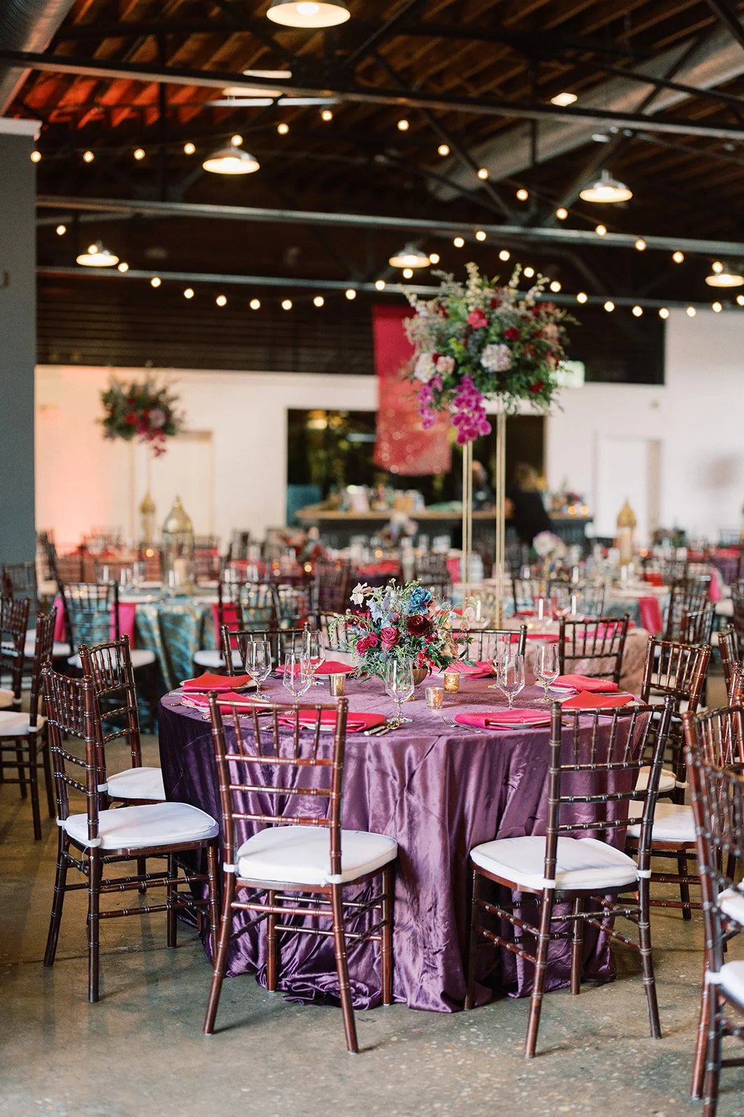A decorated event space with round tables covered in purple and multi-colored tablecloths, adorned with floral centerpieces and surrounded by wooden chairs with white cushions. String lights hang from the ceiling, creating a festive atmosphere.