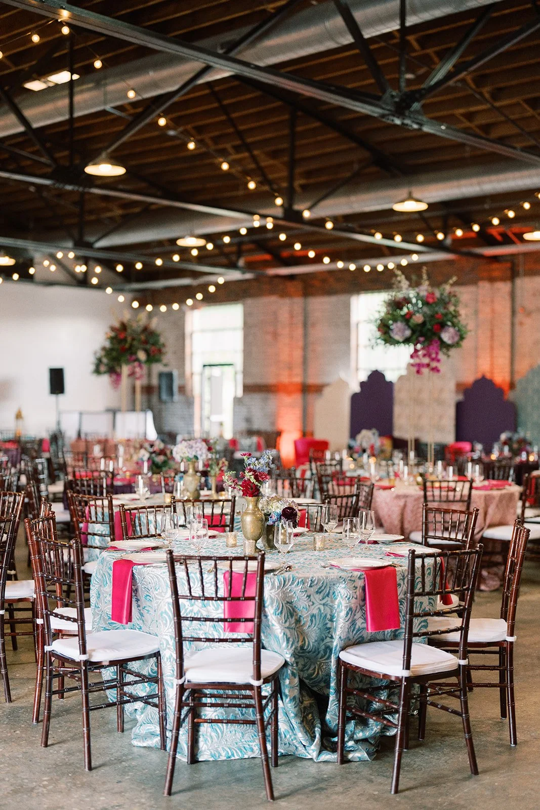 Wedding reception setup in a decorated industrial venue, featuring round tables with floral centerpieces, gold and pink accents, and hanging floral arrangements, with string lights on the ceiling.