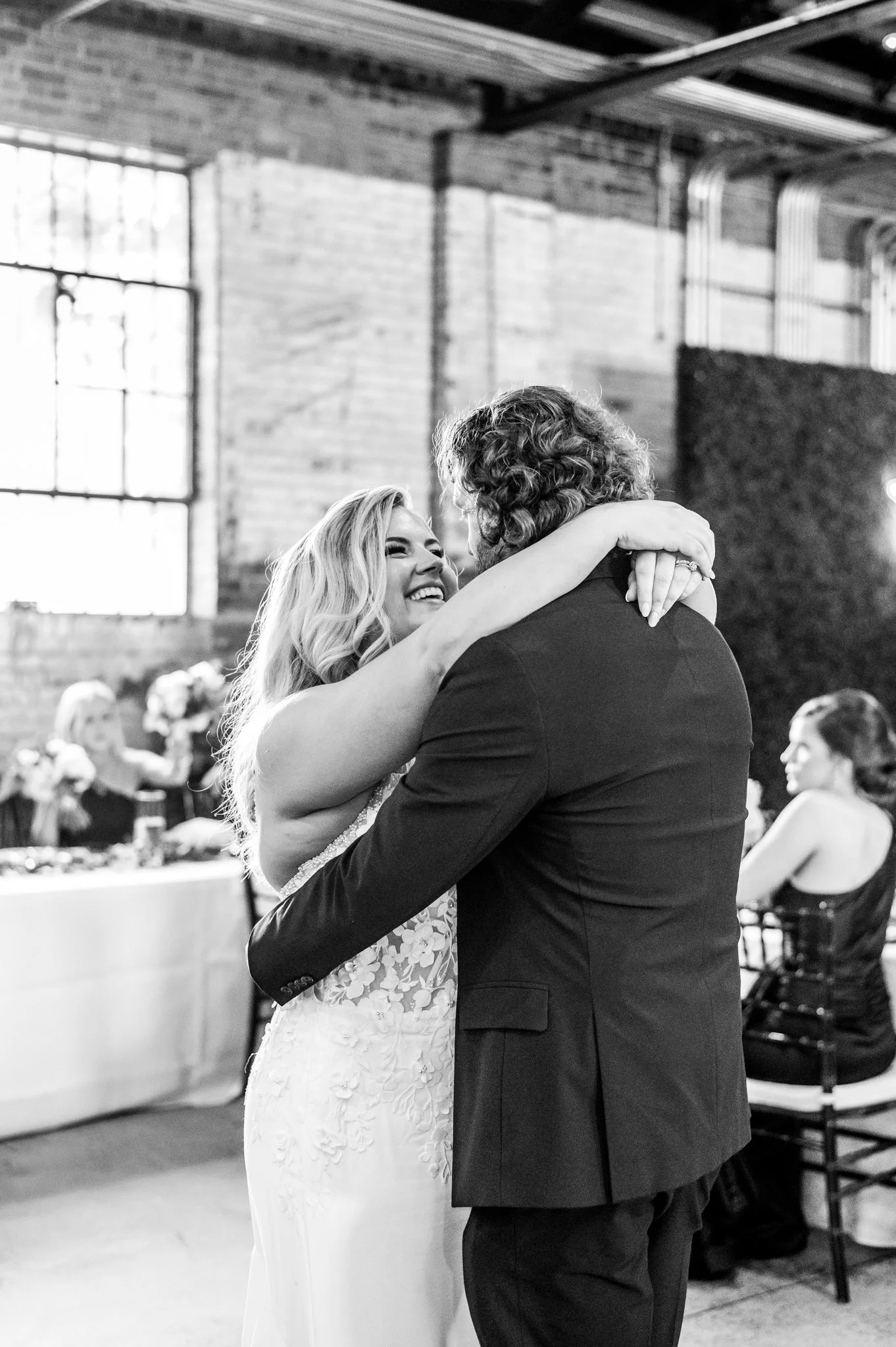 A bride and groom share a joyful dance at their wedding reception, embracing each other in a warmly lit venue with exposed brick walls.