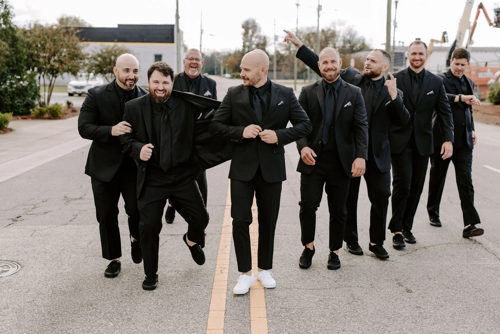 A group of seven men in black suits and black shirts walking on a street, laughing and smiling, with some pointing and one playfully pulling on the suit of the man in front. The background includes trees, a building, and construction equipment.