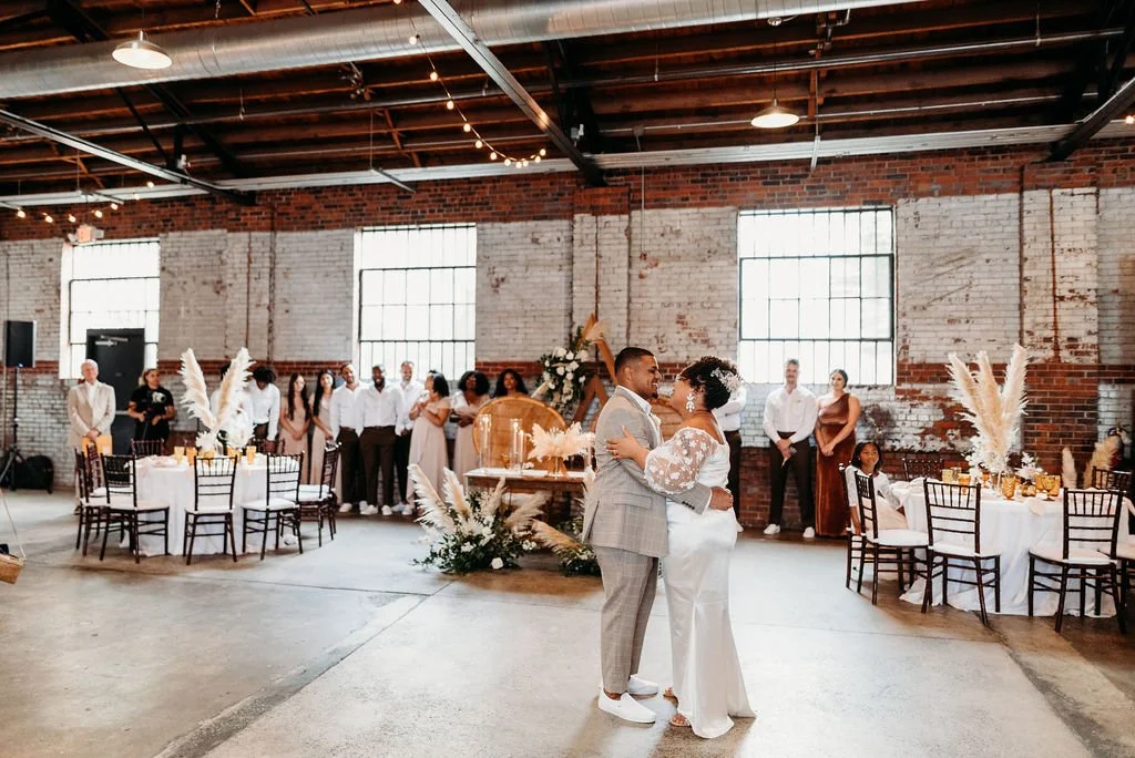 A wedding reception with a bride and groom dancing in a rustic industrial-style venue with brick walls, large windows, and string lights. Guests stand and sit around decorated tables.