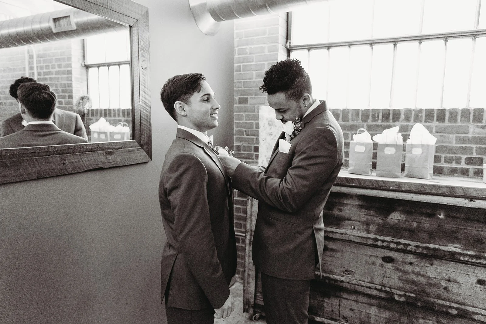 Two men in suits, one appears emotional, standing close to each other indoors with exposed brick wall and three gift bags in the background.
