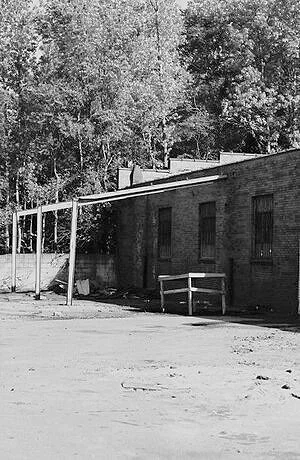 A black and white photo of an old brick building with three windows, an empty bench, and trees in the background.