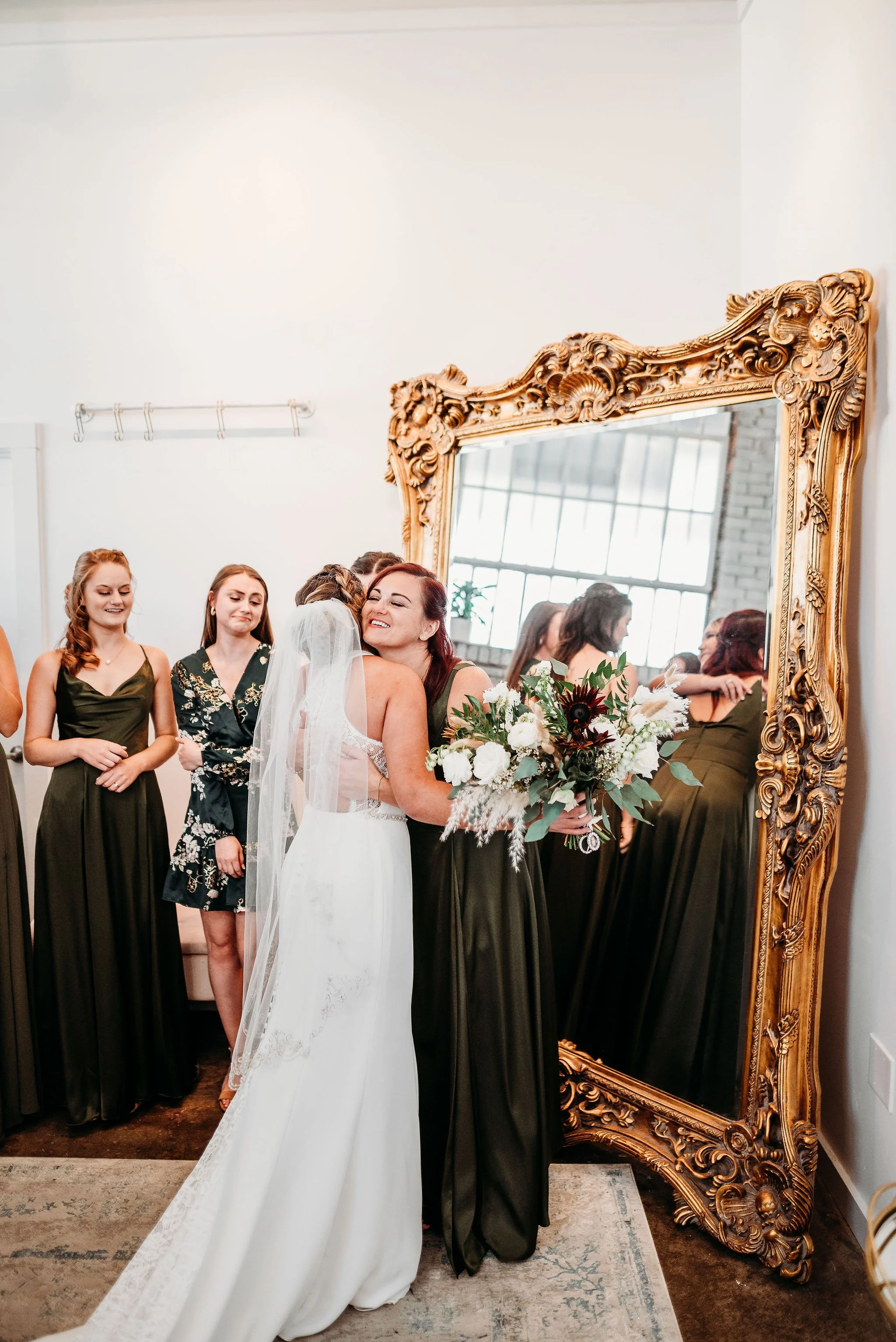A bride hugging a woman holding a bouquet of flowers in front of a large ornate mirror, surrounded by bridesmaids at a wedding celebration.