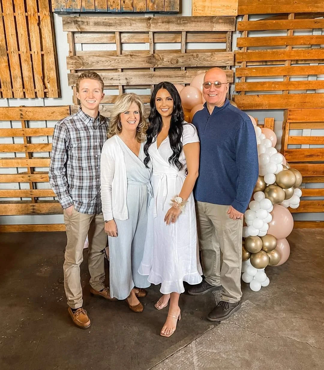 Four people, a young man, two women, and an older man, stand together smiling for a photo. They are indoors in front of a decorative wooden backdrop with balloons in beige, white, and gold.