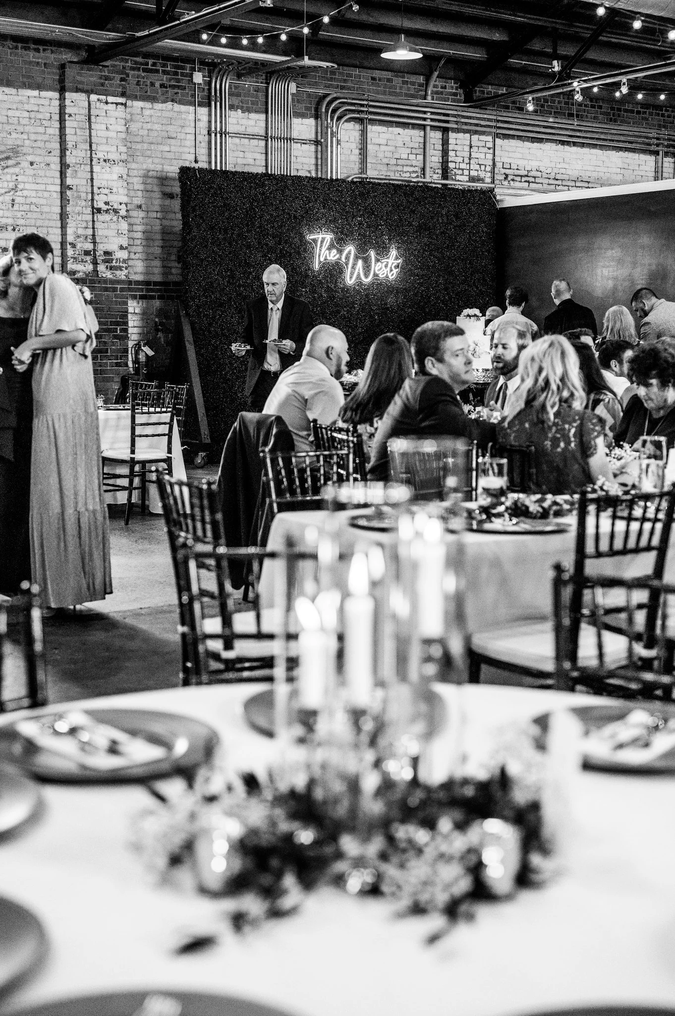 A black and white photo of a social gathering or party with people seated at tables, some standing and talking, in an industrial-style venue with exposed brick walls and visible piping. A man in a suit is standing near a decorated wall with a neon si
