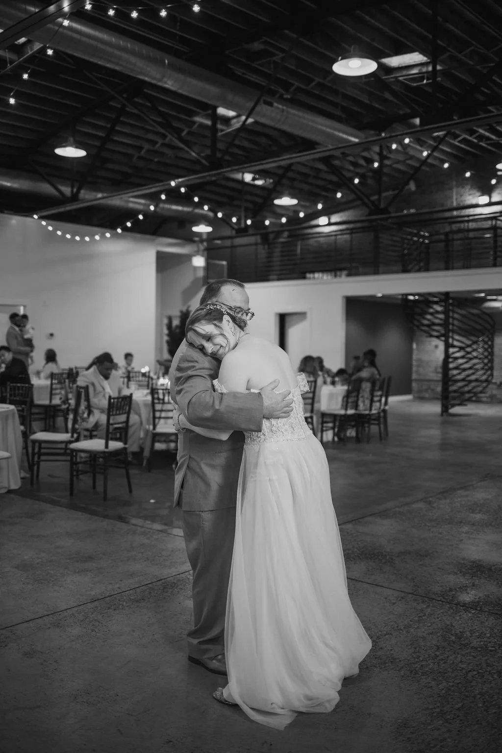 A father dancing with his daughter, possibly at her wedding reception, in a decorated indoor venue with tables and guests in the background.