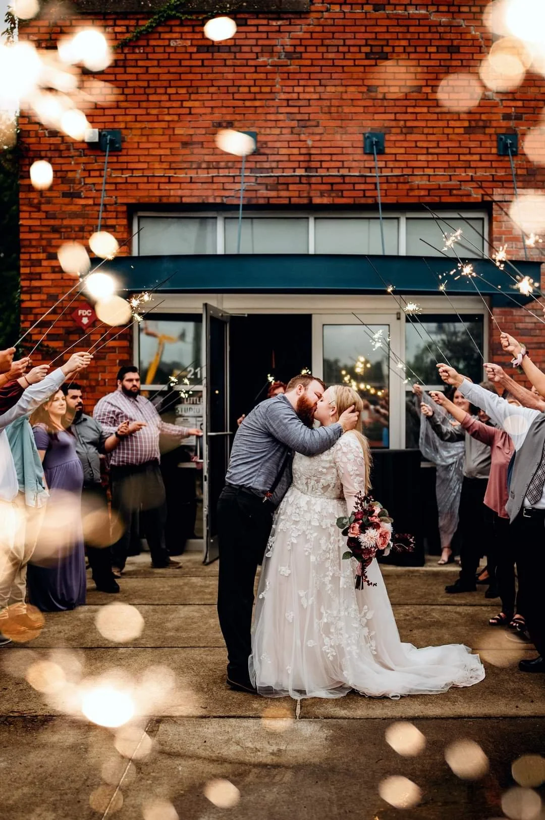 Couple sharing a kiss at their wedding celebration outside a brick building surrounded by guests holding sparklers.
