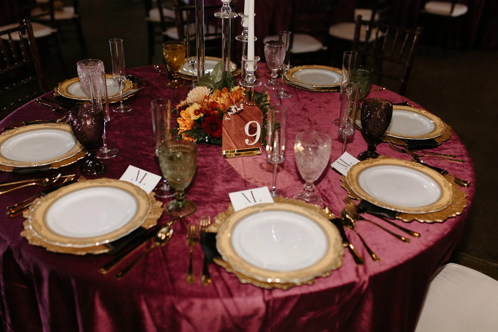 Round table set for a formal dinner with gold-rimmed white plates, gold cutlery, colored glassware, and a floral centerpiece with candles, on a pink tablecloth, with a table number card in the center.