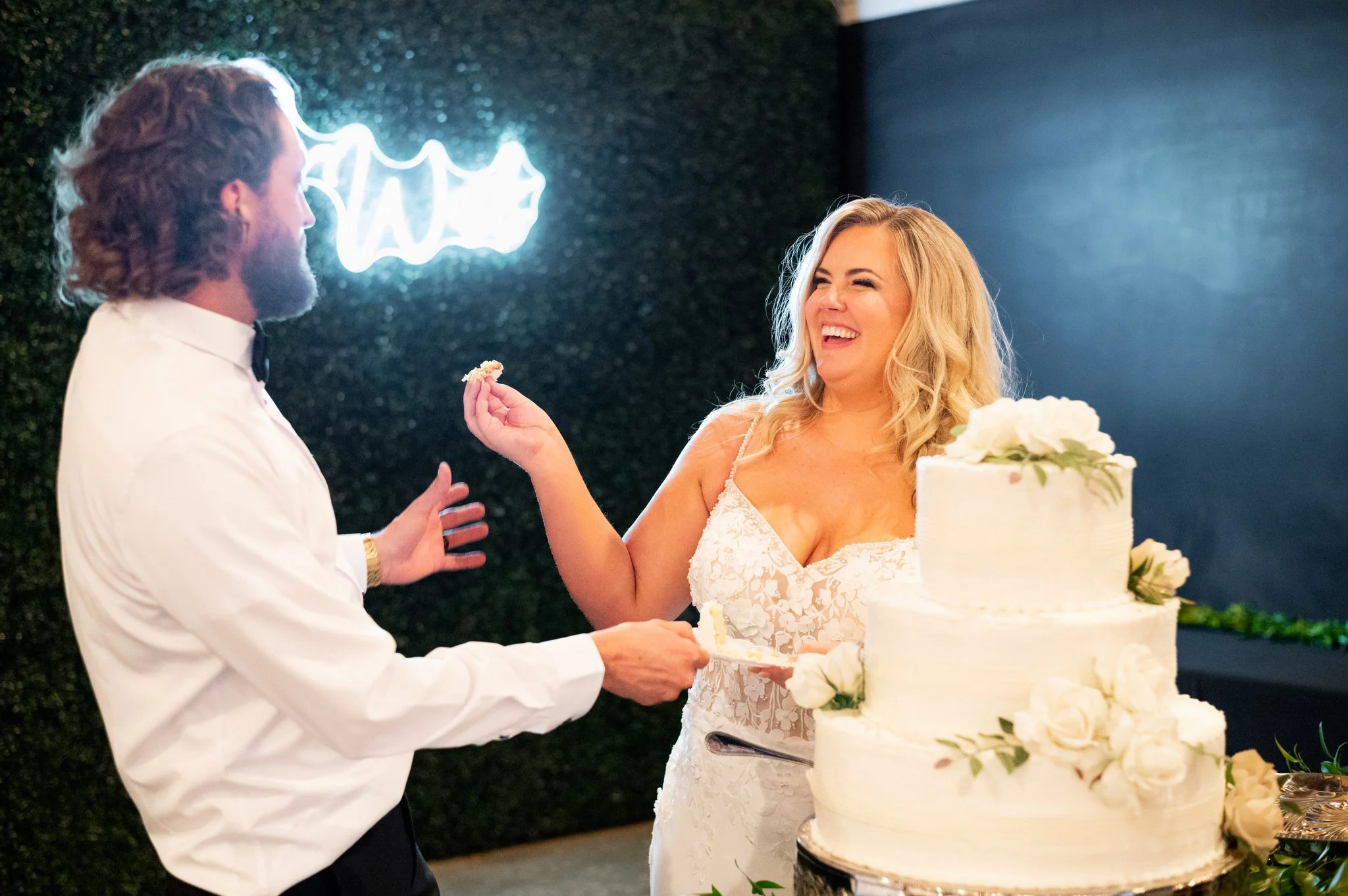 A bride and groom smiling and holding a cake knife during their wedding cake cutting ceremony, with a three-tier white wedding cake decorated with white flowers in the foreground.