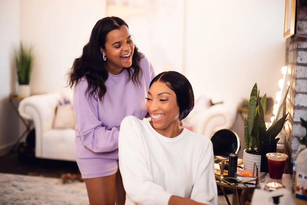 Two women are smiling and laughing in a cozy living room. One is sitting with short black hair and a white sweatshirt, the other is standing with long dark hair and a purple outfit. The room has a white sofa, plants, and decorative lights.